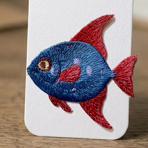 Pode incluir: Design de peixe bordado em azul e vermelho sobre fundo branco. O peixe tem um corpo azul com barbatanas e cauda vermelhas. O peixe tem uma textura bordada detalhada, com um pequeno olho e manchas.