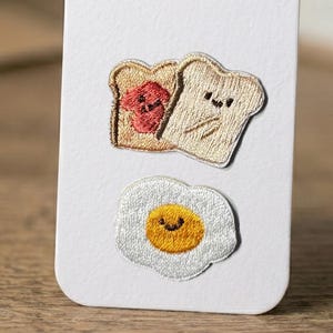 May include: Embroidered patches of breakfast food. Two slices of toast, one with jam, and a fried egg with a smiling yolk. The patches are beige, white, yellow, and red.