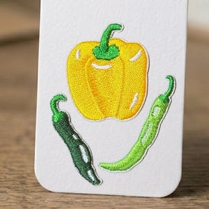 Puede incluir: Parches bordados con un pimiento amarillo y dos chiles verdes. El pimiento está centrado, con los chiles colocados debajo. Los parches están sobre un fondo blanco, mostrando los colores vibrantes y las puntadas detalladas.