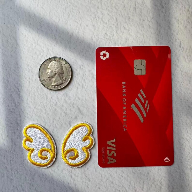 May include: A red Bank of America Visa credit card, a quarter, and two embroidered butterfly wing patches with white and yellow thread are displayed on a white surface. The card has a chip and the Visa logo.