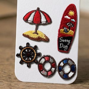 May include: Embroidered patches with a nautical theme. Features a red and white striped umbrella, a red surfboard with a sunny day scene, a ship's wheel, and two life preservers.