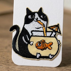 Op de afbeelding: Geborduurde patch met een zwart-witte kat die uit een vissenkom drinkt. De kat houdt een vissenkom vast met een oranje vis en een rietje. De patch heeft een gele achtergrond en een zwarte omtrek.