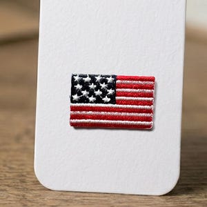 May include: A rectangular embroidered patch featuring the United States flag design. The patch displays a dark blue canton with white embroidered stars, positioned next to alternating horizontal red and white embroidered stripes.
