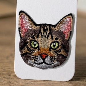 May include: Embroidered patch featuring a detailed cat face. The patch showcases a tabby cat with brown and black fur, green eyes, and a red nose. The cat's ears have pink inner details. The patch is likely for clothing or accessories.