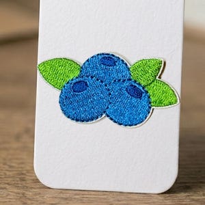 May include: Embroidered patch featuring three blue blueberries with green leaves on a white background. The blueberries have a textured appearance, and the leaves are a bright green. The patch is likely for clothing or accessories.