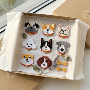 May include: A collection of nine embroidered dog patches in a gift box. The patches feature various dog breeds with detailed stitching and vibrant colors. The box includes decorative elements like dried lavender and bone-shaped patches.