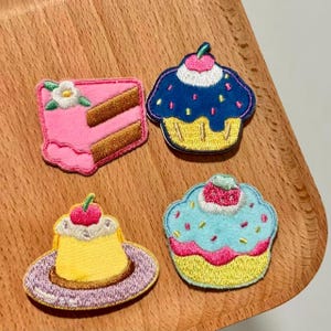May include: Four embroidered patches featuring desserts. Includes a pink cake slice with a white flower, a blue cupcake with sprinkles, a yellow custard with a cherry, and a light blue cupcake with pink frosting.