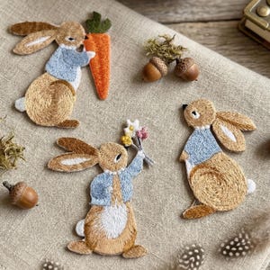 Op de afbeelding: Geborduurde konijnenpatches in bruin-, beige- en blauwtinten. Eén konijn houdt een wortel vast, een ander een boeket bloemen. De patches liggen op een neutrale linnen achtergrond, met eikels en veren.