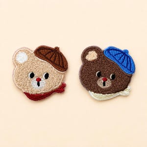 May include: Two embroidered bear patches. The left bear is light tan with a brown beret and a red scarf. The right bear is dark brown with a blue beret and a white scarf. Both bears feature black eyes, a red nose, and white muzzle details.