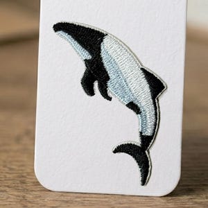 Könnte beinhalten: Gestickter Aufnäher mit einem Orca-Wal-Design. Der Aufnäher hat ein Schwarz-Weiß-Farbschema mit detaillierten Nähten. Der Orca wird in einer dynamischen Pose dargestellt, mit gebogenem Körper und ausgestreckten Flossen.