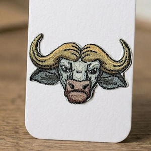 May include: Embroidered patch featuring a detailed buffalo head. The patch showcases a buffalo with tan horns, a gray face, and a brown muzzle. The design is set against a white background, highlighting the intricate stitching and color palette.