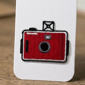 May include: A red camera embroidered patch featuring a black lens, a silver flash unit on top, and a white border. The rectangular design includes a small silver square detail near the lens and black accents on the sides, all stitched onto a white card.