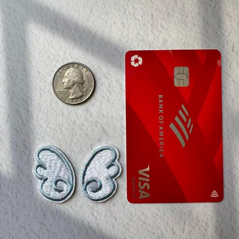 May include: A red Bank of America Visa credit card with a silver logo and chip, a silver quarter, and two small, light blue embroidered wing patches. The patches have a detailed, feathery design.