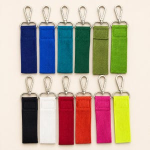 May include: A collection of twelve rectangular fabric keychains arranged in two rows. Each keychain features a woven strap in a distinct solid color, including blue, green, black, white, red, orange, pink, and yellow, attached to a silver metal lobster clasp.