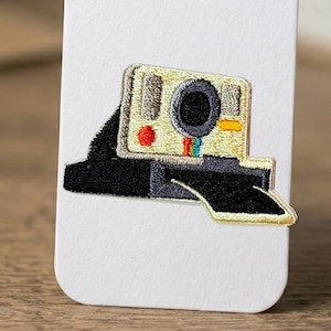 May include: Embroidered patch featuring a vintage Polaroid camera design. The patch has a beige body with a black lens and base. It includes a red button and a rainbow stripe detail, perfect for adding to clothing or accessories.