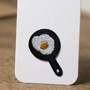 May include: Embroidered patch featuring a fried egg in a black frying pan. The egg has a white yolk and a golden yellow yolk. The patch is attached to a white card.