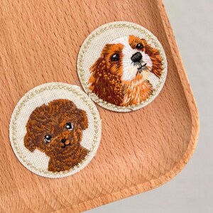 May include: Two embroidered dog patches. One patch features a brown and white Cavalier King Charles Spaniel, and the other depicts a brown poodle. Both patches are round with a gold stitched border, set on a wooden tray.