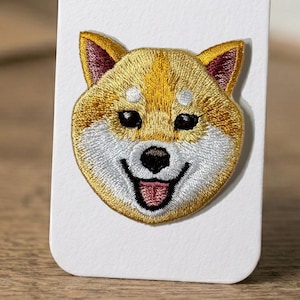 Puede incluir: Parche bordado con la cara de un perro Shiba Inu. El parche tiene un esquema de color amarillo y blanco con ojos negros y una lengua rosa. Las orejas del perro son de un tono rojo más oscuro. El parche está sobre un fondo blanco.