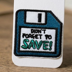 May include: Embroidered patch in the shape of a teal floppy disk with black trim. The patch has the text "DIDN'T FORGET TO SAVE!" in black and green. The patch is attached to a white surface.