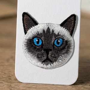 Pode incluir: Patch bordado com o rosto de um gato siamês. O gato tem um rosto branco e cinza com olhos azuis brilhantes e orelhas marrons escuras. Para roupas ou acessórios.