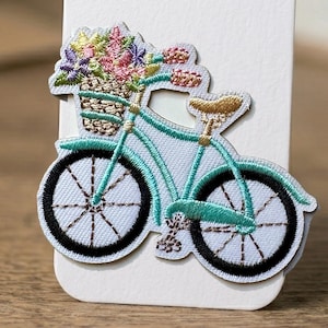 May include: Embroidered patch of a light blue bicycle with a basket of colorful flowers. The bicycle has black wheels and teal accents. The patch is on a white background.