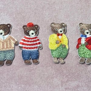 Op de afbeelding: Vier geborduurde berenpatches. Elke beer is bruin met een wit gezicht en draagt verschillende outfits: een trui, een gestreept shirt, een gele jas en een rode jas. De patches staan op een lichtpaarse achtergrond.