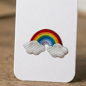 May include: An embroidered patch featuring a colorful rainbow arching over two white clouds. The rainbow consists of red, orange, yellow, green, blue, and purple thread. The clouds are stitched with white thread and have a textured appearance.
