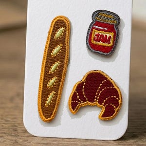 May include: Three embroidered patches on a white card: a long brown baguette with light green diagonal slashes, a small red jar with a yellow label reading JAM in red letters, and a dark red crescent-shaped croissant with golden yellow edges.