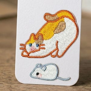 May include: Embroidered patch featuring a playful cat and a mouse. The cat is depicted in orange, white, and brown, with blue eyes and whiskers. The mouse is light gray with a pink nose. The patch is on a white background.