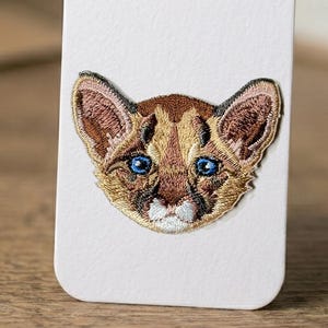 Op de afbeelding: Geborduurde patch met het gezicht van een poema-jong. De patch heeft een gedetailleerd ontwerp met bruin, beige en goudkleurig garen. De poema heeft blauwe ogen en roze binnenoren. De patch staat op een witte achtergrond.