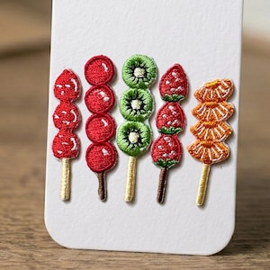 May include: Embroidered food skewers featuring strawberries, kiwi, and other fruits. The skewers are red, green, and orange, with detailed stitching and wooden sticks. The design is on a white background.