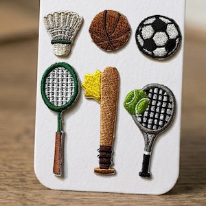 May include: A set of six embroidered sports patches on a white card. The patches include a white shuttlecock, a brown basketball, a black and white football, a green tennis racket, a brown baseball bat with a yellow star, and a tennis racket with a green ball.