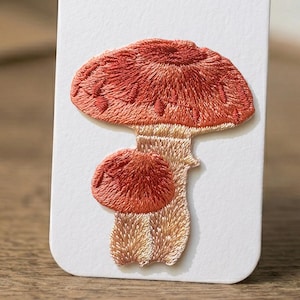 Könnte beinhalten: Gestickter Aufnäher mit einem detaillierten Pilzdesign. Der Pilz hat einen rot-orangen Hut und einen beigefarbenen Stiel mit aufwendigen Stickereien. Der Aufnäher befindet sich auf einem weißen Hintergrund, der die Textur und Farbe der Stickerei hervorhebt.