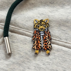 May include: Embroidered patch of a leopard wearing a leopard print coat. The patch is in yellow, orange, and black. It is set against a light grey fabric. A green drawstring with a silver tip is in the upper left corner of the image.