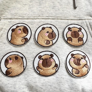 May include: Six embroidered patches featuring a cartoon capybara design. Each patch has a white border and depicts a capybara with brown fur, pink cheeks, and a dark brown nose. The patches are arranged on a light gray fabric.