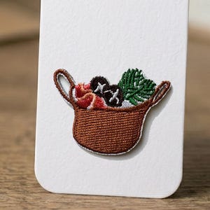 Peut inclure: Écusson brodé représentant un panier marron rempli d'objets rouges, noirs et verts. Le panier a deux anses et est posé sur un fond blanc. L'écusson est probablement destiné aux vêtements ou aux accessoires.