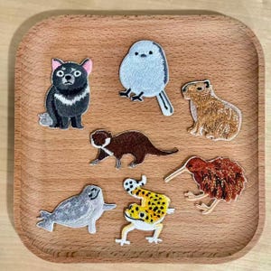 Op de afbeelding: Een verzameling geborduurde dierenpatches, waaronder een Tasmaanse duivel, een vogel, een capibara, een otter, een kiwi, een zeehond en een geel-zwart gevlekte kikker. De patches zijn gerangschikt op een houten dienblad.