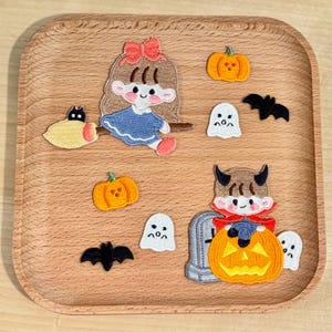 May include: A collection of Halloween-themed patches on a wooden tray. The patches include a witch riding a broom, pumpkins, ghosts, bats, and a devil character. The patches are colorful and feature cartoon-style designs.