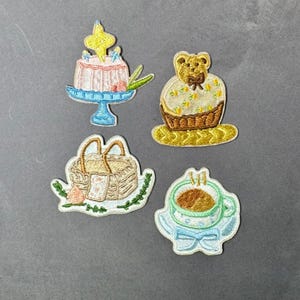 Peut inclure: Quatre écussons brodés représentant un gâteau sur présentoir, un cupcake en forme d'ours, un panier de pique-nique et une tasse de café. Les écussons sont de couleurs pastel avec des détails fins, parfaits pour la personnalisation.