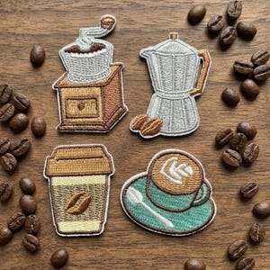 Op de afbeelding: Geborduurde patches met koffiethema's: een koffiemolen, een espressomaker, een koffiebeker voor onderweg en een koffiekopje met latte art. Koffiebonen zijn verspreid rond de patches op een houten oppervlak.