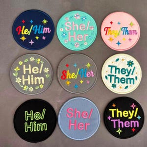 May include: Nine embroidered patches with gender pronouns. The patches are round and feature text like "He/Him", "She/Her", and "They/Them". Each patch has a different color background, including navy, teal, pink, gray, and black, with colorful embroidered accents.