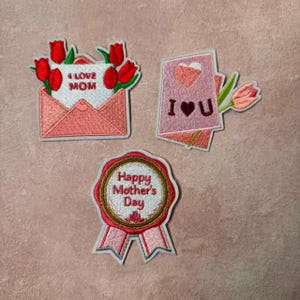 May include: Embroidered patches with a Mother's Day theme. One patch features an envelope with red tulips and the text "I LOVE MOM." Another shows a card with a heart and the text "I U." The third patch is a badge with the text "Happy Mother's Day."