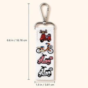 Peut inclure: Un porte-cl&eacute;s en tissu blanc avec des illustrations brod&eacute;es d'un scooter rouge, d'un v&eacute;lo orange, d'un scooter rose et d'un scooter blanc. Il comprend un mousqueton en m&eacute;tal argent&eacute; et mesure 16,76 cm de long et 3,81 cm de large.