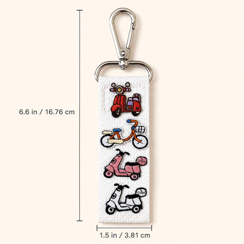 Peut inclure: Un porte-cl&eacute;s en tissu blanc avec des illustrations brod&eacute;es d'un scooter rouge, d'un v&eacute;lo orange, d'un scooter rose et d'un scooter blanc. Il comprend un mousqueton en m&eacute;tal argent&eacute; et mesure 16,76 cm de long et 3,81 cm de large.