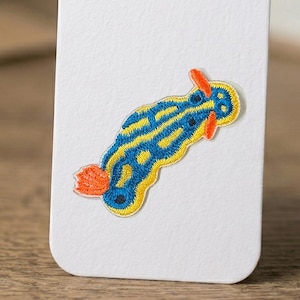 May include: Embroidered patch featuring a colorful sea slug design. The patch has a blue body with yellow stripes, orange accents, and a yellow border. The patch is attached to a white surface.
