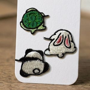 Puede incluir: Tres parches bordados con diseños de animales: una tortuga verde con caparazón texturizado, un conejo blanco con orejas de interior rosa y un panda blanco y negro. Los parches están dispuestos sobre una tarjeta rectangular blanca de esquinas redondeadas.