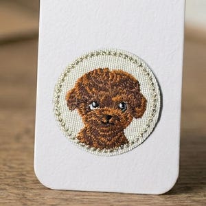 Può includere: Toppa ricamata raffigurante un cane barboncino marrone con un aspetto dettagliato e strutturato. Il muso del cane è centrato su uno sfondo circolare bianco sporco con un bordo cucito color oro. La toppa è fissata su una superficie bianca.
