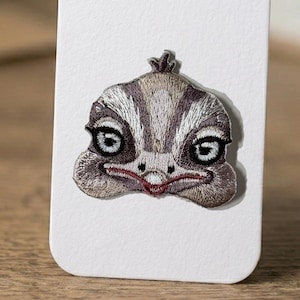 May include: Embroidered patch featuring an ostrich face. The patch has a detailed design with shades of gray, brown, and white, with a red beak and dark eyes. The patch is likely for clothing or accessories.