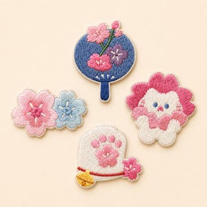 May include: Four embroidered fabric patches featuring a circular blue fan with pink and purple flowers, a cluster of pink and light blue blossoms, a white creature with pink hair, and a white cat paw design with a small yellow bell and pink flower.