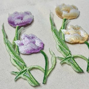 May include: Embroidered tulip patches in shades of purple and yellow with green stems and leaves. The floral patches are on a light beige fabric. These patches can be used for clothing or craft projects.